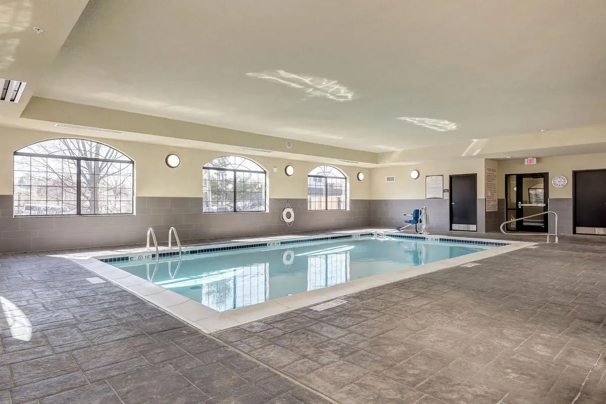 The image displays an inviting indoor pool area, featuring a sparkling pool surrounded by large windows that let in natural light. Poolside seating encourages relaxation and socializing, with a calm atmosphere that signals comfort and leisure. The scene is well-maintained and welcoming, suggesting a year-round retreat for swimming and unwinding.