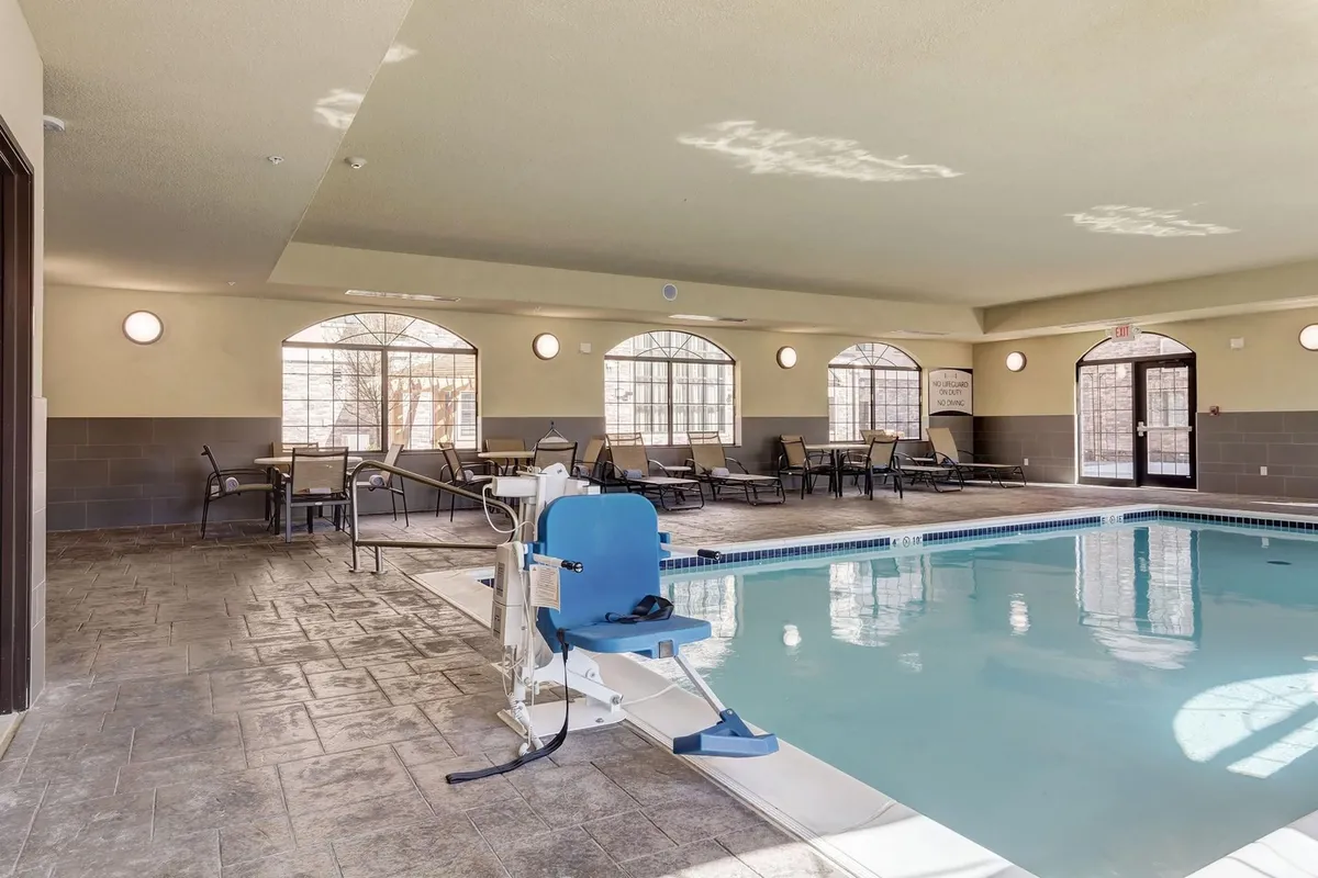 A serene indoor swimming pool with large windows allowing natural light and a wheelchair lift for accessibility. The pool area is equipped with seating for relaxation or socializing, emphasizing inclusive design and leisure opportunities.