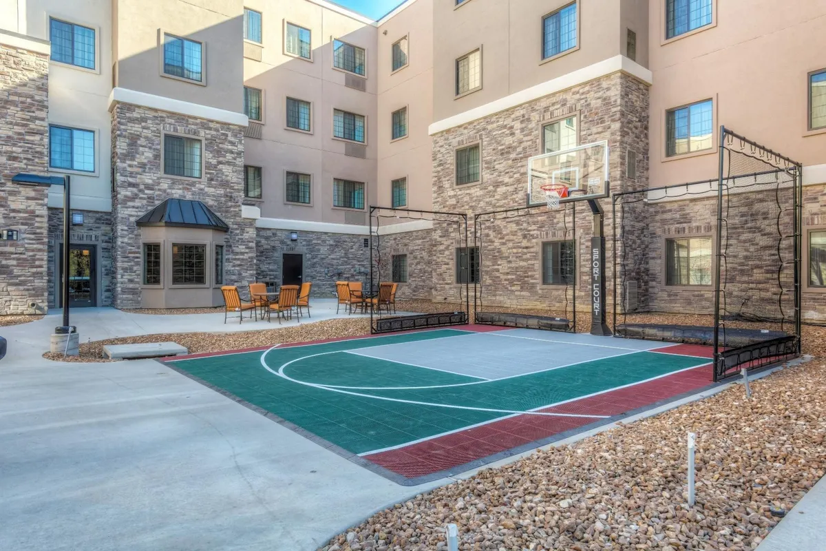 This exterior image shows the Staybridge Suites St. Louis – Westport's building facade, a lively basketball court, and an inviting outdoor seating area. People are seen playing basketball and socializing in the sun, highlighting the hotel's active community feel and connection to the surrounding business hub.