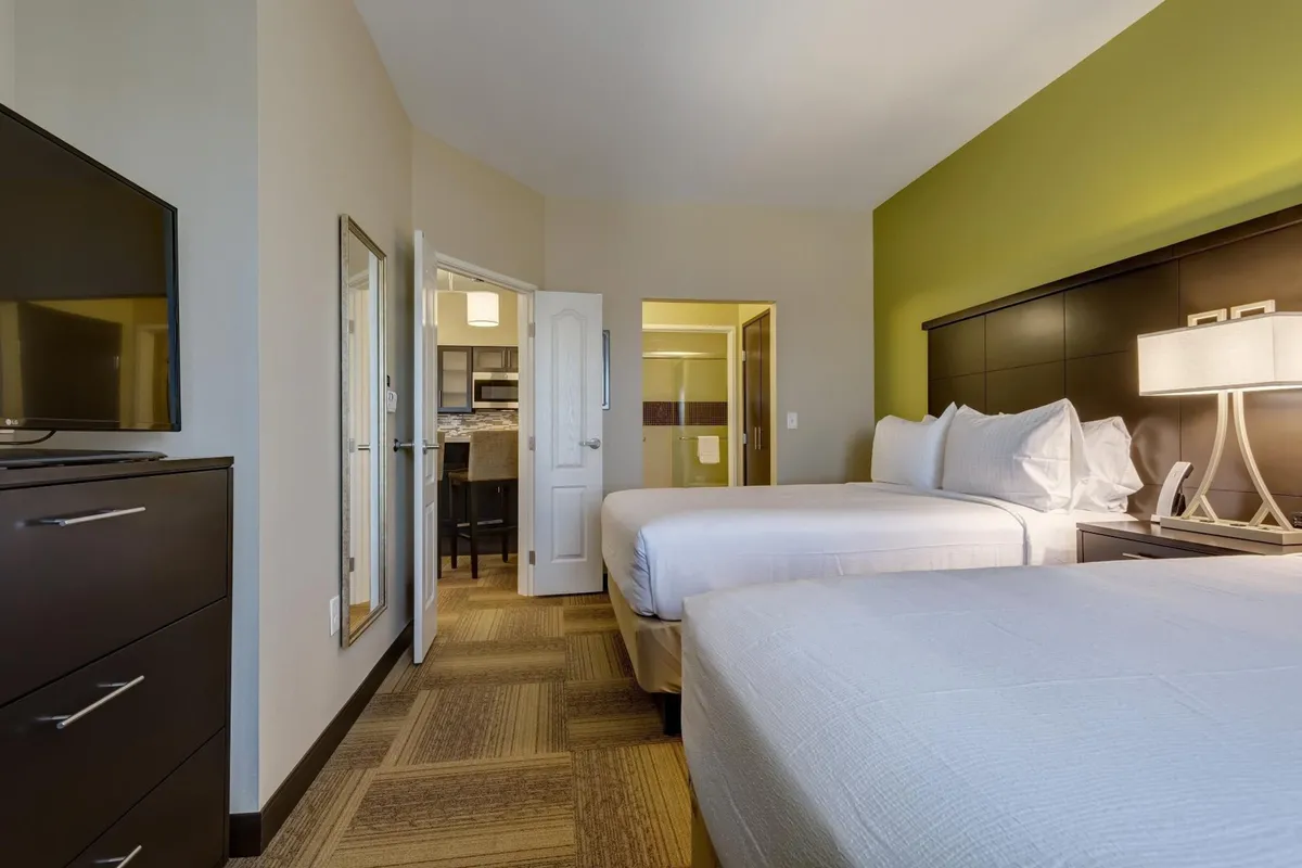 A suite interior featuring two queen beds with plush linens, a TV opposite the beds, a dedicated desk area, and a doorway leading to a kitchen. The decor is inviting and comfortable, suggesting a home-away-from-home vibe ideal for families or business travelers.