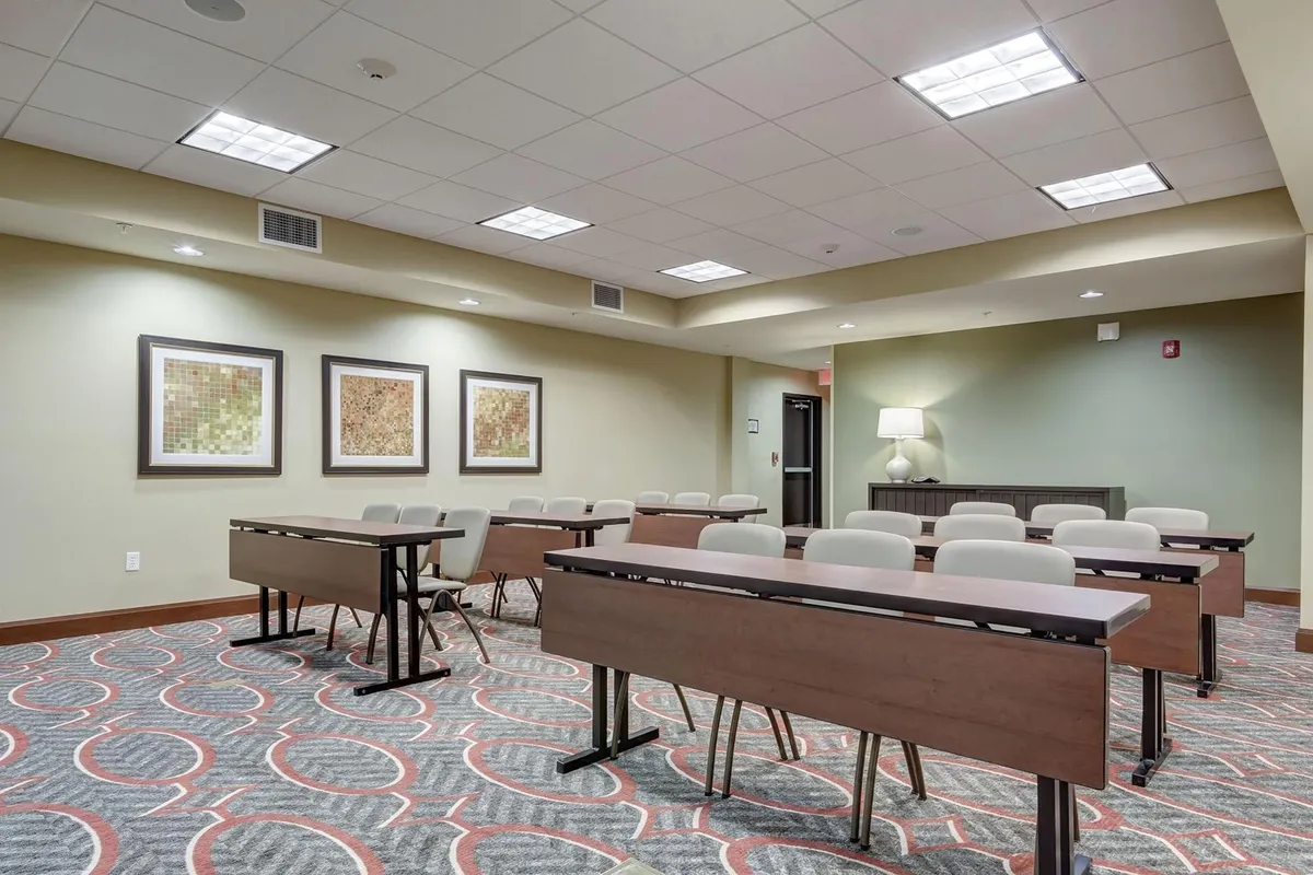 This image features a collaborative meeting space with rectangular tables surrounded by upholstered chairs, accented by framed artwork and a desk lamp. The setup is clean, modern, and designed to foster interaction or productivity, with an inviting, well-lit atmosphere.