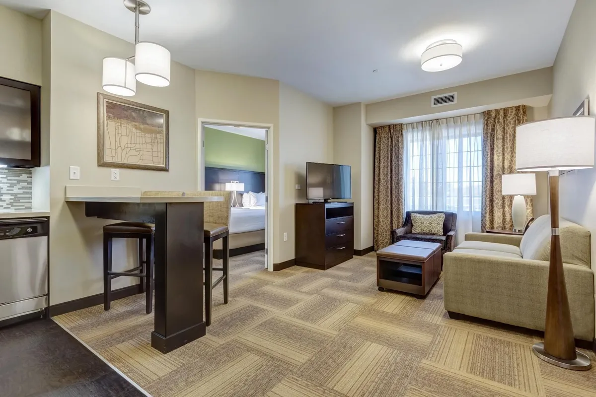 The image showcases a warm, inviting living area within the suite, featuring a modern bar-height table with stools perfect for meals or gathering, a plush sofa and armchair for relaxing, soft natural light streaming through curtained windows, and a sleek TV unit for entertainment.