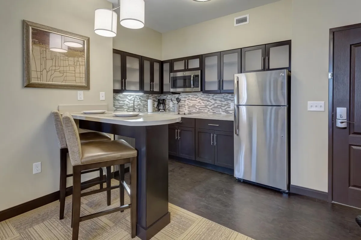 A cozy living area with a bar counter, stools, kitchen appliances, decorative wall art, and inviting lighting. The scene reflects a welcoming, multi-functional space that can be used for relaxing, casual dining, or socializing.