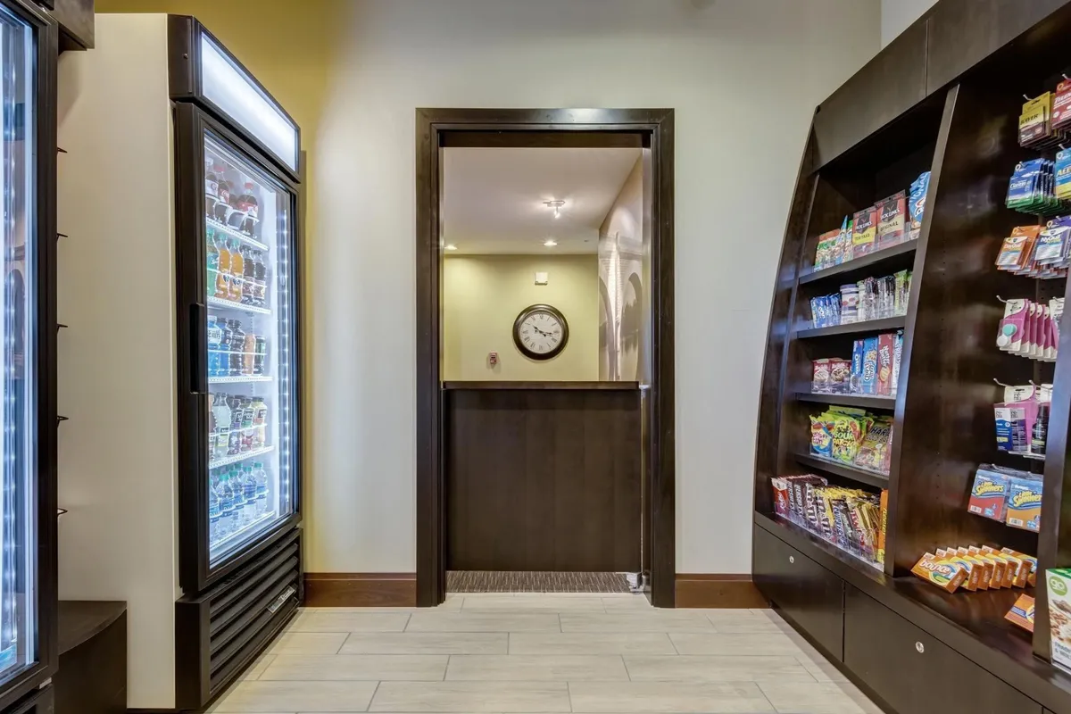 A convenient snack and drink area with a refrigerated display and neatly arranged snack options, set against a backdrop of a clean, casual interior. A clock on the wall suggests accessibility and ease for guests needing quick refreshments.