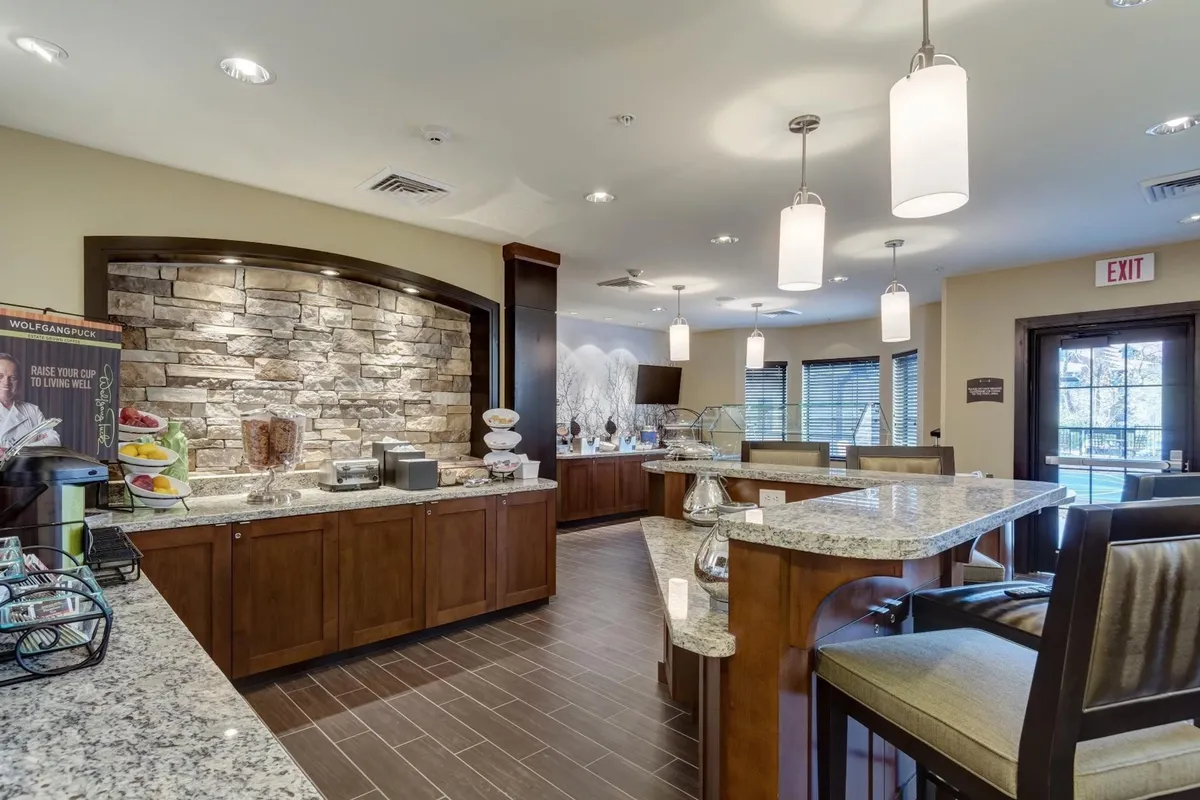 The image showcases a granite countertop, stone wall, a cozy seating area, and a buffet display. Guests are seen socializing over breakfast and coffee, creating a relaxed, homelike atmosphere. The presence of a buffet aligns directly with the complimentary breakfast aspect mentioned in the section.