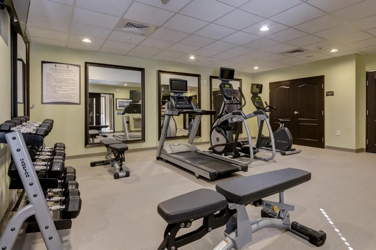 A well-lit fitness room equipped with dumbbells, a treadmill, an elliptical machine, and large mirrors, offering space for solo or group exercise routines. The atmosphere feels inviting and health-focused, reflecting the opportunity for physical wellness throughout a stay.